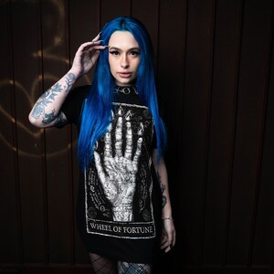 May include: A woman with blue hair is wearing a black t-shirt with a white graphic of a hand and the text "Wheel of Fortune".