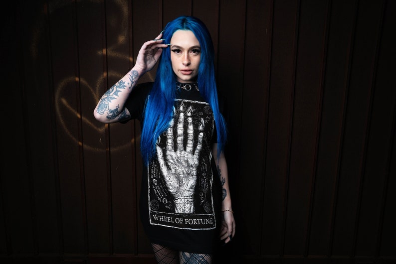 May include: A woman with blue hair is wearing a black t-shirt with a white graphic of a hand and the text "Wheel of Fortune".