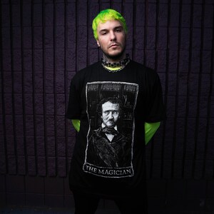 May include: A man with green hair is wearing a black t-shirt with a white image of Edgar Allan Poe and the text "The Magician".