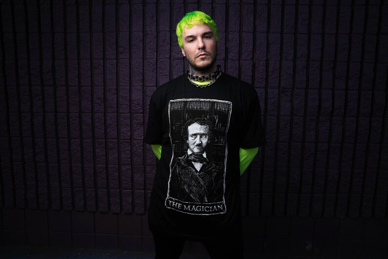 May include: A man with green hair is wearing a black t-shirt with a white image of Edgar Allan Poe and the text "The Magician".