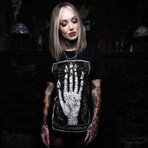May include: Black t-shirt featuring a white graphic of a hand with flames and the text "Wheel of Fortune". The model is wearing the shirt and has tattoos on both arms.