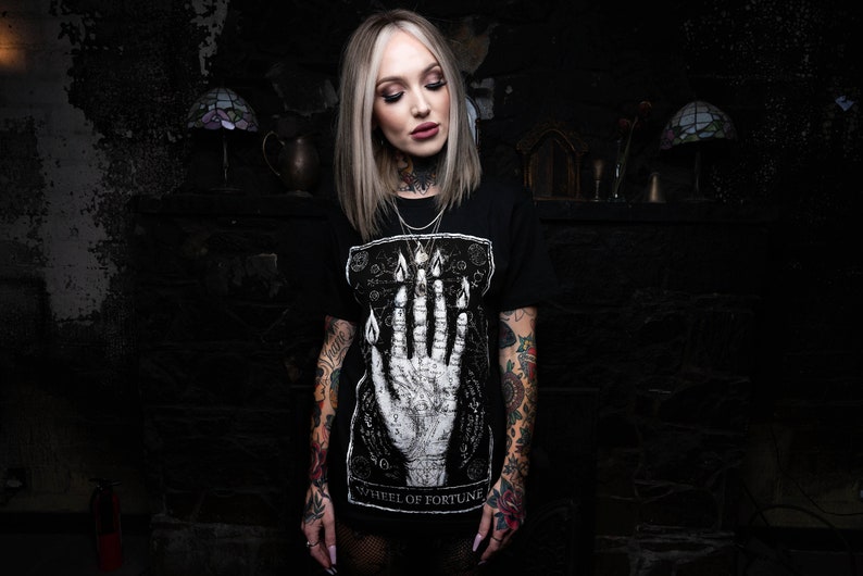 May include: Black t-shirt featuring a white graphic of a hand with flames and the text "Wheel of Fortune". The model is wearing the shirt and has tattoos on both arms.