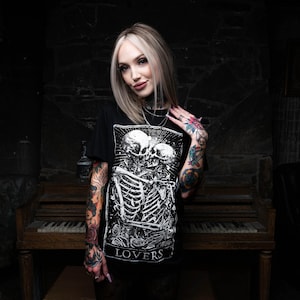 May include: A woman with blonde hair and tattoos is wearing a black t-shirt with a white graphic of two skeletons embracing. The graphic includes the word "LOVERS".