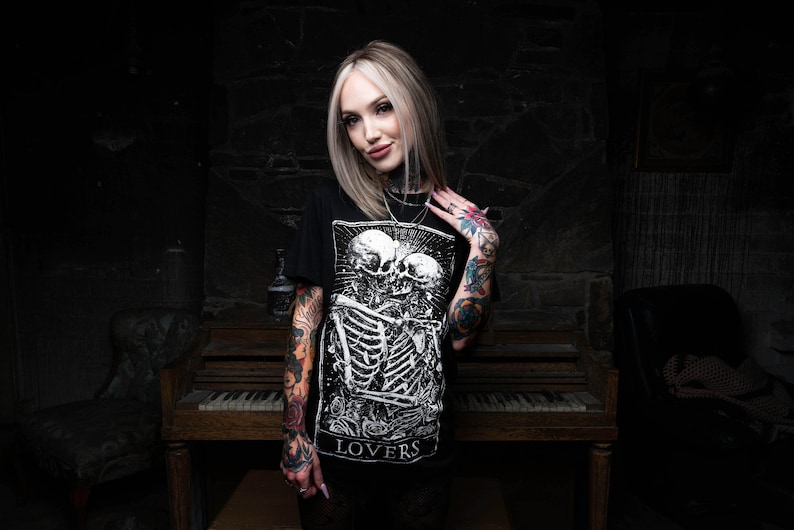 May include: A woman with blonde hair and tattoos is wearing a black t-shirt with a white graphic of two skeletons embracing. The graphic includes the word "LOVERS".