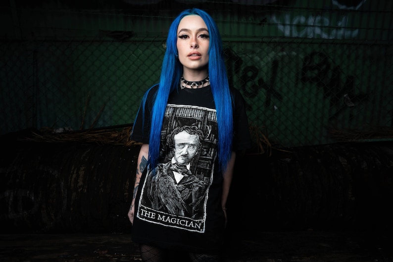 May include: A woman with bright blue hair is wearing a black t-shirt with a white graphic of a man with a raven on his shoulder. The graphic is labelled "The Magician".