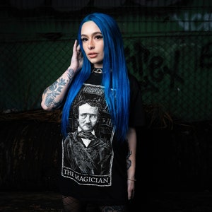 May include: A woman with bright blue hair is wearing a black t-shirt with a white graphic of a man with a raven on his shoulder. The graphic is labelled "The Magician".