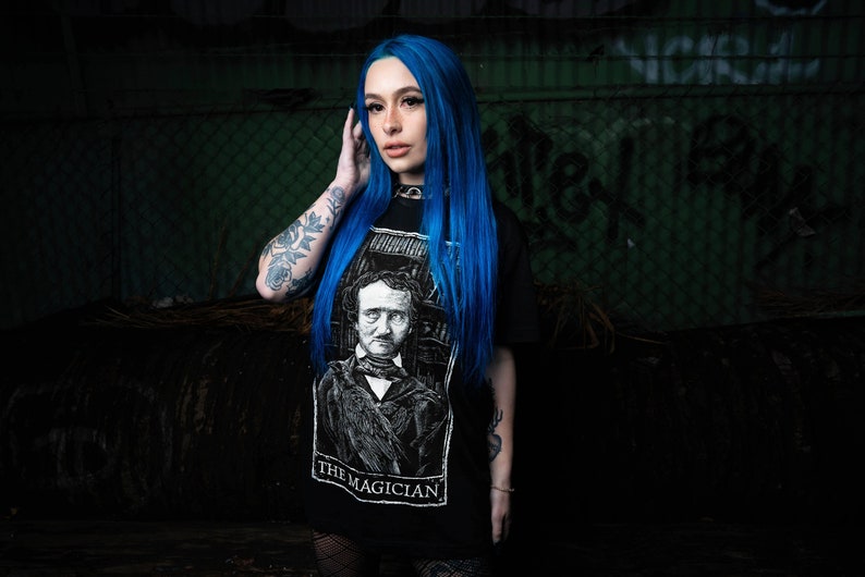 May include: A woman with bright blue hair is wearing a black t-shirt with a white graphic of a man with a raven on his shoulder. The graphic is labelled "The Magician".