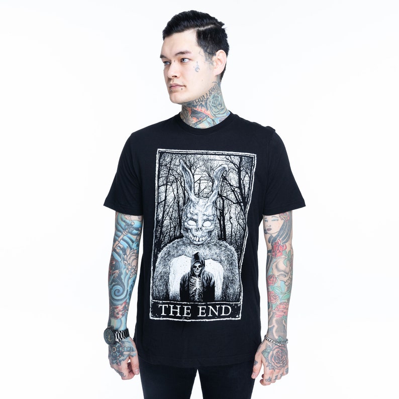 K&ouml;nnte beinhalten: Schwarzes T-Shirt mit einem wei&szlig;en Grafikdruck eines Kaninchens mit Maske und einer Sense. Die Grafik ist in einem Tarotkarten-Rahmen mit dem Text "THE END" dargestellt.