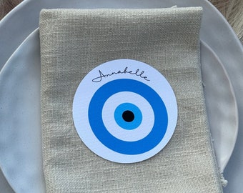 May include: A white napkin with a fringe border is folded on a white plate. A round paper place card with a blue and black evil eye design and the name "Annabelle" is on top of the napkin.