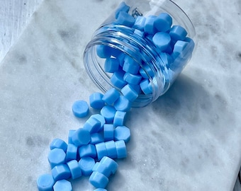 May include: A clear plastic jar tipped over, spilling light blue wax seal beads onto a white and gray marble surface. The hexagonal beads are scattered around the jar, creating a visually appealing arrangement. Ideal for crafts and stationery.