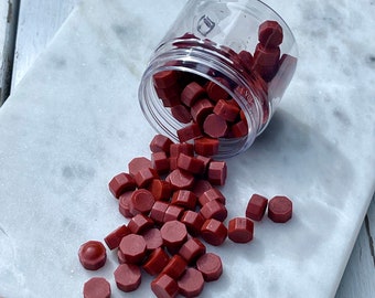 May include: A clear plastic jar filled with red sealing wax pellets, spilling out onto a white marble surface. The wax pellets are hexagonal in shape.