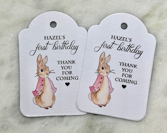 May include: Two white favor tags with scalloped edges. Each tag features a cartoon illustration of a rabbit wearing a pink jacket. The text on the tags reads "Hazel's first birthday" and "Thank you for coming".