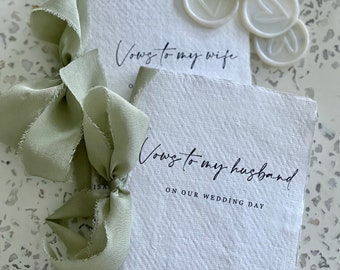 Puede incluir: Dos libros encuadernados en papel blanco con el texto "Vows to my wife" y "Vows to my husband on our wedding day" escrito en tinta negra. Los libros están atados con una cinta verde salvia y hay cuatro sellos de cera blanca con un diseño de corazón en la parte superior del libro.