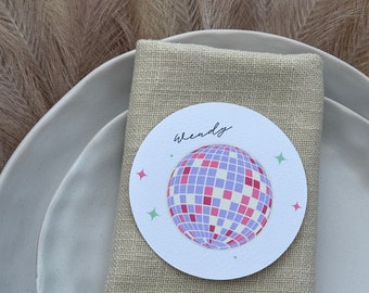 May include: A white round place card with a purple and pink disco ball graphic and the name "Wendy" written in black cursive. The place card is on top of a beige linen napkin.
