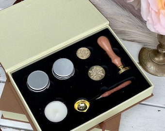 May include: A set of wax seal supplies in a beige box with a brown lid. The set includes a wooden handle, a metal spoon, a candle, two wax sticks, and two wax seals. The box is open and the supplies are visible.