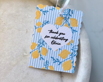 May include: A decorative gift tag with a scalloped edge, featuring a blue and white striped background with yellow lemons and blue leaves. The tag has a white heart with the text "Thank you for celebrating Olivia". A white ribbon is attached.