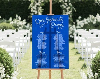 May include: A blue sign on a wooden easel with the words "Our Favourite People" in white script. The sign lists names under the headings "one," "two," "three," and "four." The background shows white chairs set up for an outdoor event.