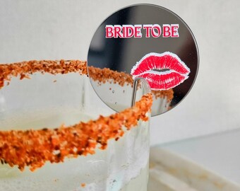 May include: A circular silver mirror drink garnish featuring the red text BRIDE TO BE and a graphic of red lips. The garnish is attached to the rim of a glass filled with a light-colored beverage and a chili-powder-coated rim.