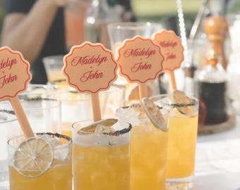 May include: A set of light peach-colored drink stirrers with a scalloped circular top, each featuring the red printed text 'Madelyn + John' in a cursive script. These stirrers are placed inside clear glasses filled with an orange liquid and ice.