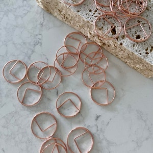 Könnte beinhalten: Ein Satz roségoldener Büroklammern mit geometrischem Design. Die Klammern sind in einem Kreis auf einer weißen Marmorfläche angeordnet.