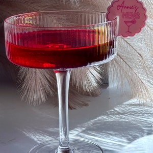 May include: A clear glass cocktail glass filled with a red liquid. A pink paper cocktail stirrer with the text "Annie's Hens" is attached to the rim of the glass.
