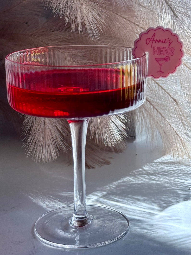 May include: A clear glass cocktail glass filled with a red liquid. A pink paper cocktail stirrer with the text "Annie's Hens" is attached to the rim of the glass.