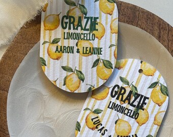 May include: Two oval-shaped gift tags with a lemon and leaf pattern. The tags have the words "GRAZIE LIMONCELLO" in green and black text, with names below. A white ribbon is attached to one tag. The tags are on a white and wooden surface.