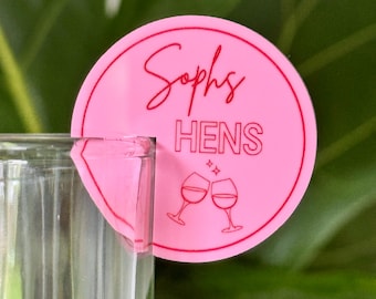 May include: A pink drinks charm with the text "Sophs HENS" in a script font, and two wine glasses. The charm is attached to a clear glass. The background is a green leafy plant.
