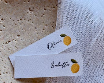 May include: Two rectangular white paper place cards featuring a printed yellow lemon illustration with a green leaf. The top card displays the name Olivia in black script, and the bottom card displays the name Isabella in black script.