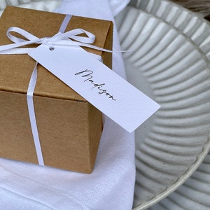 K&ouml;nnte beinhalten: Eine kleine braune Geschenkbox mit einem wei&szlig;en Band und einem wei&szlig;en Geschenkanh&auml;nger mit der Aufschrift "Madison". Die Box liegt auf einer wei&szlig;en Serviette und einem wei&szlig;en und grauen Teller.