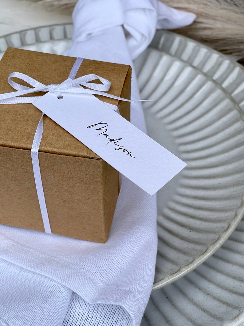 K&ouml;nnte beinhalten: Eine kleine braune Geschenkbox mit einem wei&szlig;en Band und einem wei&szlig;en Geschenkanh&auml;nger mit der Aufschrift "Madison". Die Box liegt auf einer wei&szlig;en Serviette und einem wei&szlig;en und grauen Teller.