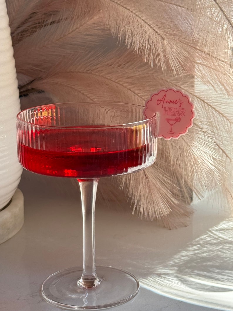 May include: A clear glass with a red drink and a pink paper tag that says "Annie's Hens" with a cocktail glass icon.