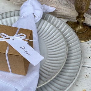 K&ouml;nnte beinhalten: Eine wei&szlig;e Serviette, die mit einem Knoten gebunden ist, eine kleine braune Geschenkbox mit einem wei&szlig;en Band und einem wei&szlig;en Geschenkanh&auml;nger mit der Aufschrift "Madison", sowie zwei wei&szlig;e Teller mit geripptem Design.