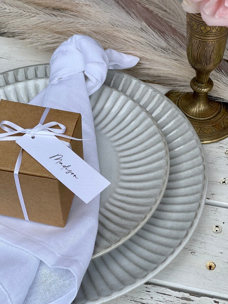 K&ouml;nnte beinhalten: Eine wei&szlig;e Serviette, die mit einem Knoten gebunden ist, eine kleine braune Geschenkbox mit einem wei&szlig;en Band und einem wei&szlig;en Geschenkanh&auml;nger mit der Aufschrift "Madison", sowie zwei wei&szlig;e Teller mit geripptem Design.