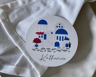 May include: White round place card with a blue and white illustration of a Greek building with a red flower pot and the name Katherine written in cursive.