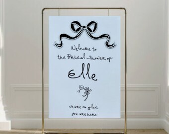 May include: A white sign with black text and a black bow on a gold stand. The sign reads "Welcome to the Bridal Shower of Elle. We are so glad you are here."