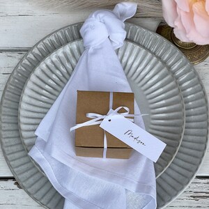K&ouml;nnte beinhalten: Eine wei&szlig;e Leinen-Serviette, die mit einem Knoten gebunden ist, liegt auf einem grauen und wei&szlig;en Keramikteller. Auf der Serviette liegt eine kleine braune Geschenkbox mit einem wei&szlig;en Etikett, auf dem "Madison" steht.