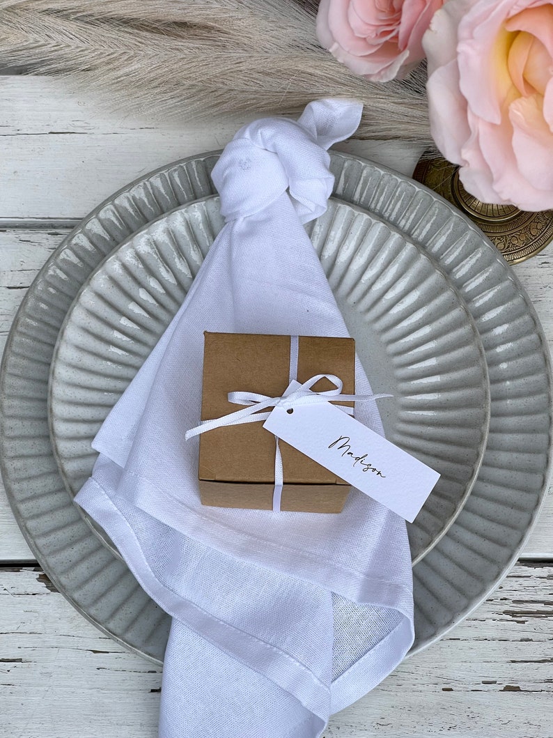 K&ouml;nnte beinhalten: Eine wei&szlig;e Leinen-Serviette, die mit einem Knoten gebunden ist, liegt auf einem grauen und wei&szlig;en Keramikteller. Auf der Serviette liegt eine kleine braune Geschenkbox mit einem wei&szlig;en Etikett, auf dem "Madison" steht.