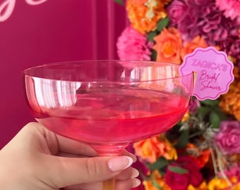 May include: A pink coupe glass filled with a pink beverage, held by a hand. A pink paper tag with the text "ZAGICA'S Bridal Shower" is attached to the glass. The background features a vibrant floral arrangement with orange, pink, and purple flowers.
