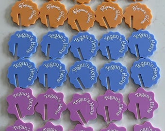 May include: A collection of colorful drink charms in orange, blue, and purple. Each charm has a unique scalloped edge and a slot for a glass stem. The text "Tegan's Hens" and "Tegan's Hena" is printed on each charm.