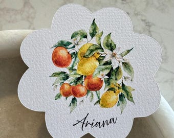 May include: A white, scalloped-edge card featuring a watercolor illustration of lemons, oranges, green leaves, and white blossoms. The name "Ariana" is written in black script below the illustration. The card is likely used for place settings or gift tags.