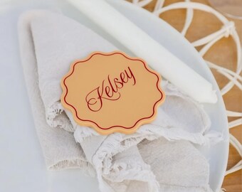 May include: A round, scalloped-edge place card in a light tan color with a thin red border and the name Kelsey written in a red cursive script, resting on a folded light beige linen napkin atop a white ceramic dinner plate.