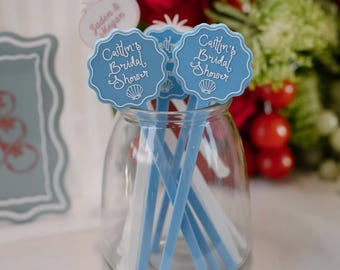 May include: A clear glass jar containing several blue and white plastic stirrers. Each stirrer features a scalloped circular top with the white text "Caitlin's Bridal Shower" and a small seashell graphic printed on the front surface.