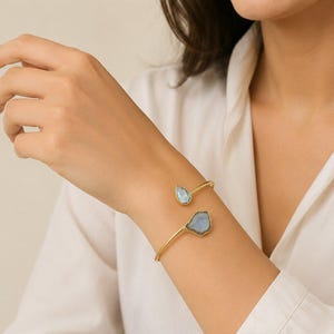 May include: A gold-toned open cuff bracelet featuring two distinct light blue stones. One stone is a small teardrop shape, while the other is a larger, irregular, faceted piece. The bracelet has a thin, polished metal band that wraps around the wrist.