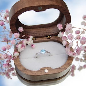 May include: A wooden ring box with a light blue gemstone ring inside. The box is open and the ring is sitting on a white cushion. The box is surrounded by pink flowers.