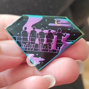 May include: A black diamond-shaped enamel pin with a holographic finish. The pin features a group of six figures standing in a line, silhouetted against a purple and blue sky with stars and a crescent moon. The figures are wearing long robes and appear to be holding hands.