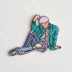 May include: An embroidered iron-on patch depicting a cartoon illustration of a person with pink hair wearing a green jacket, blue jeans, and a red and white striped tie. The person is sitting with their leg crossed and their hand on their head.