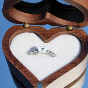 May include: A silver ring featuring a white flower-shaped accent with a small center stone, resting inside a heart-shaped wooden box with a white velvet interior. The box lid is open, and the text 5HINeeByTori is visible on the lower front.