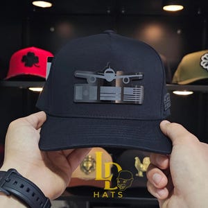 May include: A navy blue baseball cap featuring a silver airplane design above the flags of Mexico and the United States. The cap has a curved brim and a structured crown. Other hats are visible in the background.