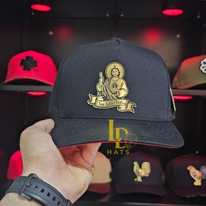 May include: Black baseball cap with a gold emblem of San Judas Tadeo. The cap has a red brim and a structured fit. Other caps with different designs are visible in the background.